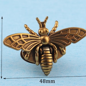 May include: A gold-toned metal bee figurine with detailed wings and a textured body. The bee is approximately 4.8 inches wide and 3.6 inches tall.