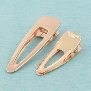 May include: Two gold metal hair clips. The clips are oval shaped with a textured edge. The clips are closed.