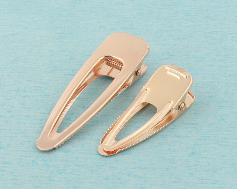 May include: Two gold metal hair clips. The clips are oval shaped with a textured edge. The clips are closed.