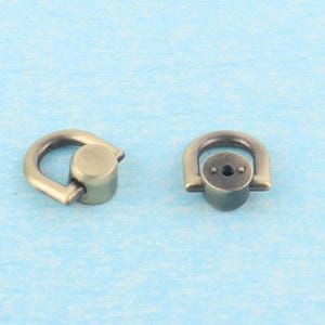 May include: Two antique brass-colored D-ring hardware pieces. Each piece has a rounded D-shape with a cylindrical base. The hardware is designed for attaching straps or other accessories to bags or other items.