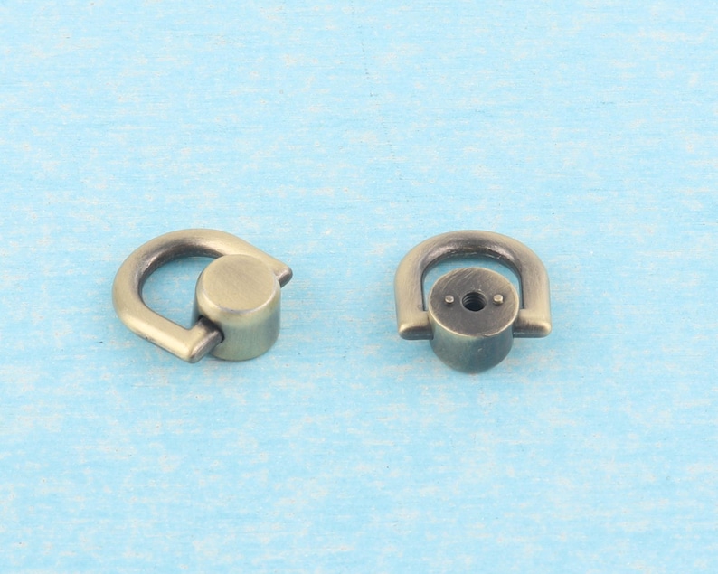 May include: Two antique brass-colored D-ring hardware pieces. Each piece has a rounded D-shape with a cylindrical base. The hardware is designed for attaching straps or other accessories to bags or other items.