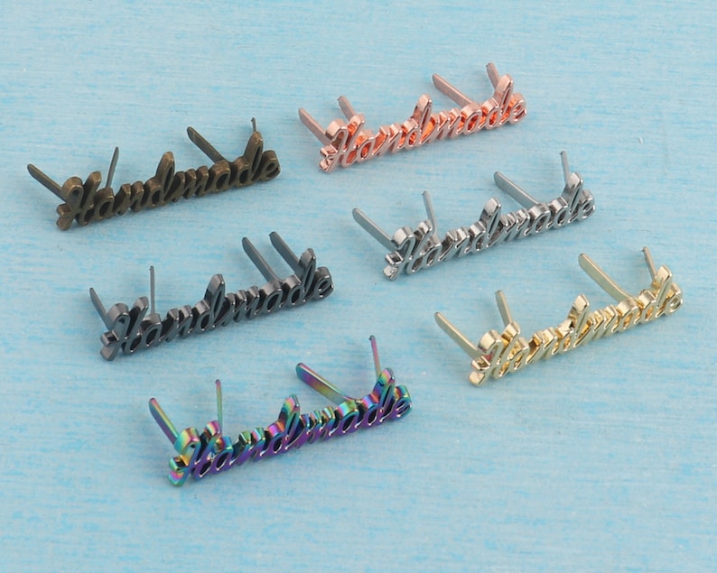 Op de afbeelding: Zes metalen "Handmade" labels met verschillende afwerkingen, waaronder goud, zilver, brons en regenboog. Elk label heeft vier pinnen om aan stof of andere materialen te bevestigen.
