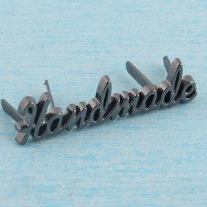Op de afbeelding: Zwart metalen label met het woord "Handmade" in een cursief schrift.