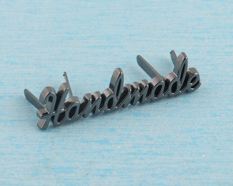 Op de afbeelding: Zwart metalen label met het woord "Handmade" in een cursief schrift.