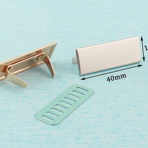 May include: Two gold-toned metal clasps with a rectangular shape and a spring mechanism. A separate piece of metal with a grid pattern is also shown. The clasps measure 40 mm by 15 mm.