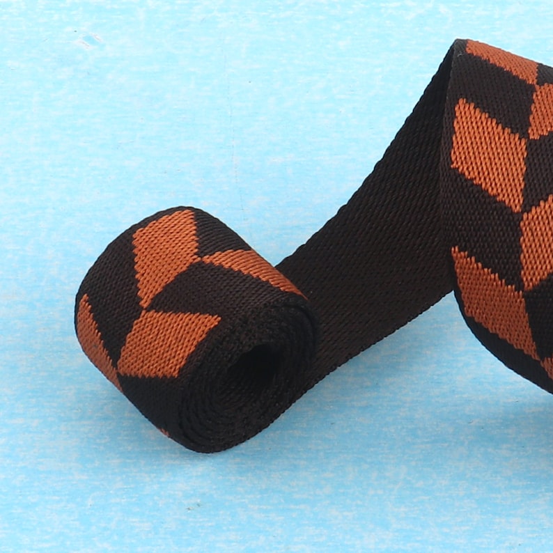 May include: Black and brown woven ribbon with a geometric pattern. The ribbon is rolled up and the pattern is visible on both sides.