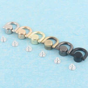 May include: An assortment of metal D-ring hardware in various finishes: silver, gold, brass, bronze, gunmetal, and black. Each D-ring is paired with a silver rivet for attachment. The hardware is displayed on a light blue surface.