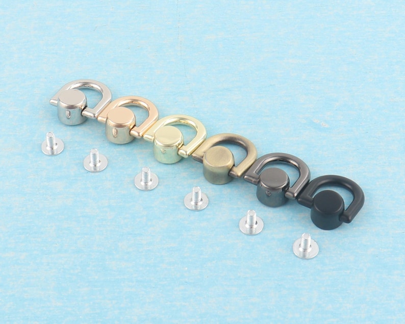 May include: An assortment of metal D-ring hardware in various finishes: silver, gold, brass, bronze, gunmetal, and black. Each D-ring is paired with a silver rivet for attachment. The hardware is displayed on a light blue surface.