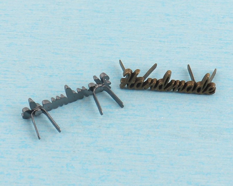 Op de afbeelding: Twee metalen labels met het woord "Handmade" erop. Het label links is zwart en het label rechts is goud.