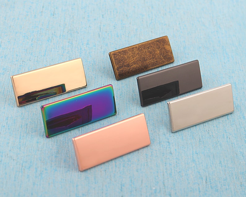 May include: Six rectangular metal name tags in different finishes: gold, rainbow, rose gold, black, silver, and antique brass.