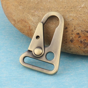 Puede incluir: Un mosquet&oacute;n giratorio de metal de color bronce, con un cuerpo triangular y una puerta con resorte. El mosquet&oacute;n tiene una parte superior redondeada y un lazo rectangular en la parte inferior. Dise&ntilde;ado para sujetar a correas u otros art&iacute;culos.
