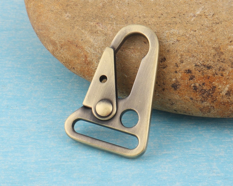 Puede incluir: Un mosquet&oacute;n giratorio de metal de color bronce, con un cuerpo triangular y una puerta con resorte. El mosquet&oacute;n tiene una parte superior redondeada y un lazo rectangular en la parte inferior. Dise&ntilde;ado para sujetar a correas u otros art&iacute;culos.