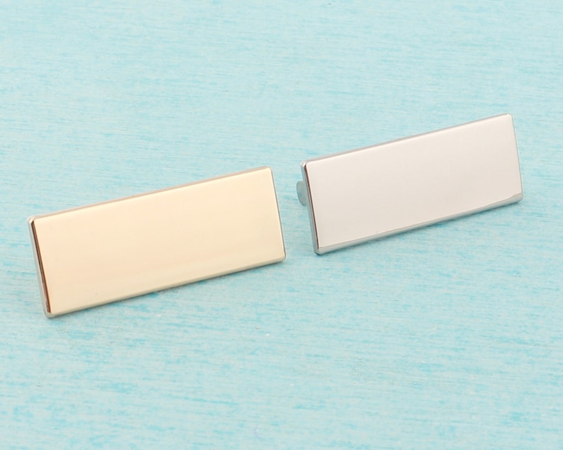 May include: Two rectangular metal name tags, one gold and one silver, on a light blue background.
