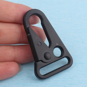Puede incluir: Un mosquet&oacute;n giratorio de pl&aacute;stico negro con un dise&ntilde;o triangular. El gancho tiene una puerta con resorte y una abertura circular para la fijaci&oacute;n. El gancho se muestra sobre un fondo azul claro.