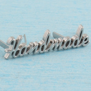 Op de afbeelding: Zilveren metalen label met het woord "Handmade" in een cursief schrift. Het label heeft twee pinnen aan de achterkant om aan een product te bevestigen.