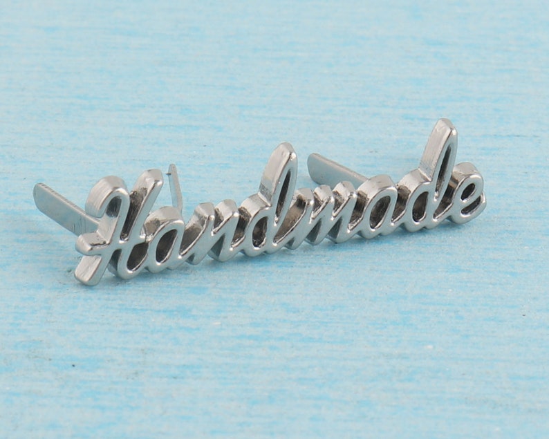 Op de afbeelding: Zilveren metalen label met het woord "Handmade" in een cursief schrift. Het label heeft twee pinnen aan de achterkant om aan een product te bevestigen.