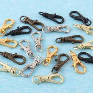 Può includere: Assortimento di moschettoni girevoli in varie finiture metalliche, tra cui oro, argento, bronzo e nero. Questi fermagli in metallo sono progettati per il fissaggio a portachiavi o cordini. I ganci sono esposti su una superficie azzurra.