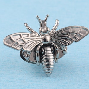 May include: A silver bee brooch with detailed wings and a textured body. The bee is perched on a small, round base.