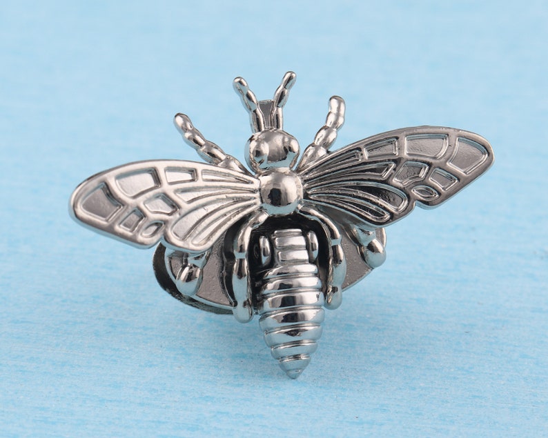 May include: A silver bee brooch with detailed wings and a textured body. The bee is perched on a small, round base.