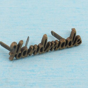 Op de afbeelding: Een goudkleurig metalen label met het woord "Handmade" erop gegraveerd. Het label heeft aan beide zijden twee kleine gaatjes om het aan een product te bevestigen.