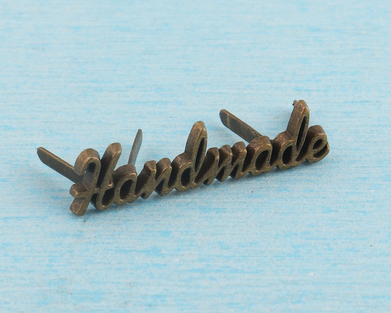 Op de afbeelding: Een goudkleurig metalen label met het woord "Handmade" erop gegraveerd. Het label heeft aan beide zijden twee kleine gaatjes om het aan een product te bevestigen.