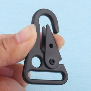 Puede incluir: Un mosquet&oacute;n de metal negro con una puerta con resorte. El mosquet&oacute;n tiene un gancho curvo en la parte superior y una abertura rectangular en la parte inferior. El clip se sostiene contra un fondo azul claro.