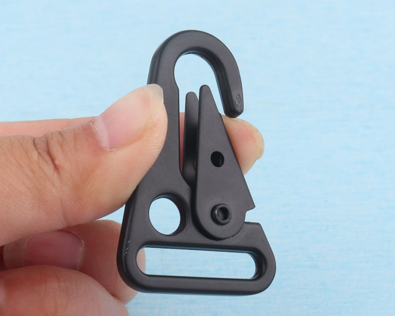 Puede incluir: Un mosquet&oacute;n de metal negro con una puerta con resorte. El mosquet&oacute;n tiene un gancho curvo en la parte superior y una abertura rectangular en la parte inferior. El clip se sostiene contra un fondo azul claro.
