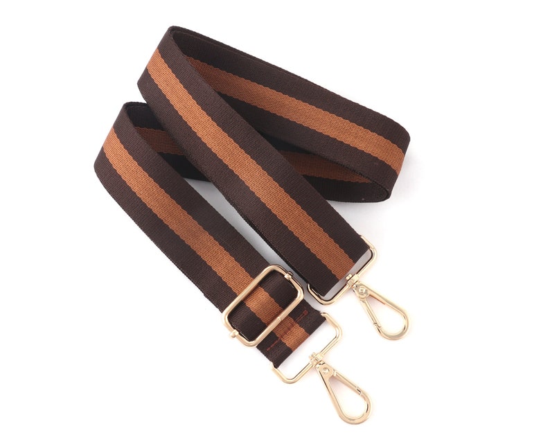 Pode incluir: Uma al&ccedil;a ajust&aacute;vel com listras marrons e bege e fechos de metal dourado. A al&ccedil;a &eacute; feita de tecido tecido e tem um visual casual.