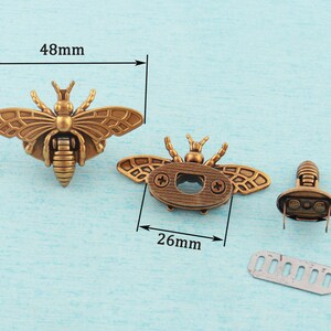 May include: Two antique gold bee-shaped metal clasps with a 48mm width and 26mm height. The clasps have a detailed design with wings, a body, and a head. One clasp has a small hole in the center of the body. The other clasp has a small metal plate attached to the back.