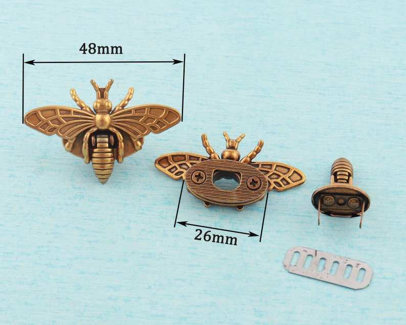 May include: Two antique gold bee-shaped metal clasps with a 48mm width and 26mm height. The clasps have a detailed design with wings, a body, and a head. One clasp has a small hole in the center of the body. The other clasp has a small metal plate attached to the back.