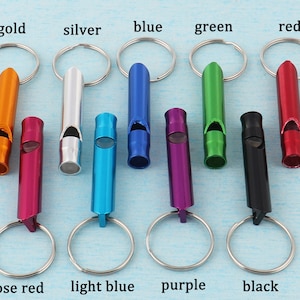 May include: A collection of colorful metal whistles with key rings. Colors include gold, silver, blue, green, red, rose red, light blue, purple, and black. The whistles are arranged in a row against a light blue background.
