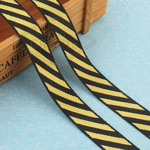 May include: Two black and yellow striped ribbons are displayed diagonally across a light blue surface. The ribbons have a repeating pattern of yellow stripes on a black background. The ribbons are likely used for crafting or decorative purposes.