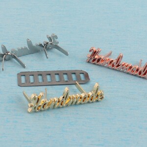Op de afbeelding: Drie metalen "Handmade" labels met verschillende afwerkingen: zilver, goud en ros&eacute;goud. Elk label heeft twee pinnen aan de achterkant om aan een product te bevestigen.