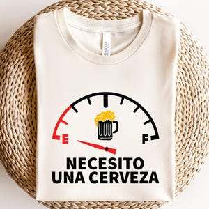 May include: A white t-shirt with a black and red fuel gauge graphic. The gauge is filled with a yellow beer mug and reads "Necesito una cerveza".