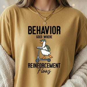 Może przedstawiać: Musztardowa koszulka z napisem "BEHAVIOR GOES WHERE REINFORCEMENT Flows". Kreskówkowa kaczka w czapce i plecaku jeździ na skuterze. Koszulka ma również ikony SVG, PNG, EPS i DXF po lewej stronie.