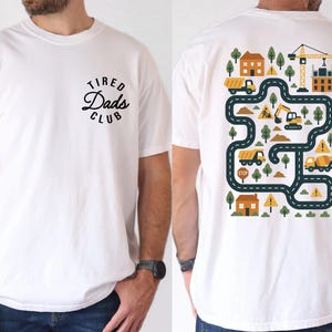 Può includere: Una t-shirt bianca con una piccola grafica di testo nera sul davanti che recita Tired Dads Club. Il retro della maglietta mostra una scena di cantiere colorata con strade, camion, escavatori, gru, case e segnali di avvertimento gialli.
