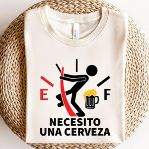 May include: A white t-shirt with a red and black fuel gauge graphic. The gauge is empty and a stick figure is holding a beer mug. The text "Necesito una cerveza" is printed below the graphic.