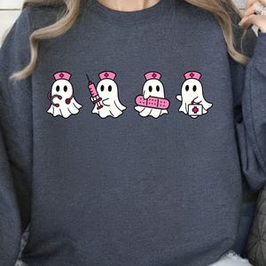 May include: A dark gray long-sleeve sweatshirt featuring a graphic of four white ghosts dressed as nurses, each wearing a pink nurse cap. The ghosts are holding medical items including a stethoscope, a syringe, a bandage, and a medical kit.