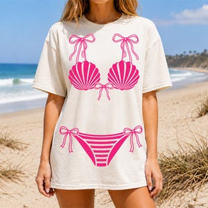 May include: A cream-colored short-sleeve t-shirt featuring a bright pink graphic print of a bikini. The design includes two seashell-shaped cups with bow-tie straps and a matching bikini bottom with side ties and horizontal stripes on the front.