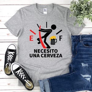 May include: A gray t-shirt with a graphic of a fuel gauge with a stick figure pushing a beer mug. The text on the shirt reads "Necesito una cerveza".