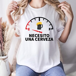 May include: White t-shirt with a black and red graphic of a fuel gauge with a beer mug icon. The gauge reads "E" for empty and "F" for full. The text below the gauge reads "Necesito una cerveza" which translates to "I need a beer".