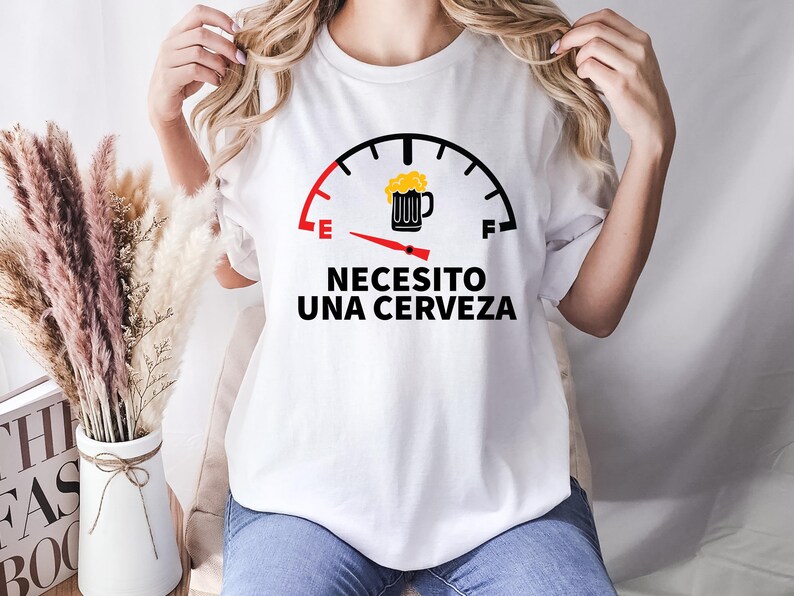 May include: White t-shirt with a black and red graphic of a fuel gauge with a beer mug icon. The gauge reads "E" for empty and "F" for full. The text below the gauge reads "Necesito una cerveza" which translates to "I need a beer".
