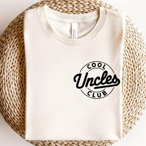 Puede incluir: Una camiseta blanca con un diseño circular negro que dice "Cool Uncles Club".