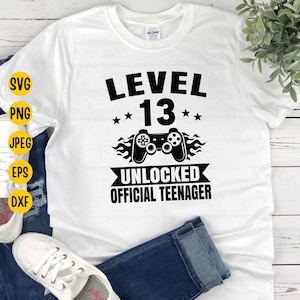 Puede incluir: Camiseta blanca con texto negro que dice "Nivel 13 Desbloqueado Adolescente Oficial". Un mando de videojuego negro con llamas está por encima del texto.