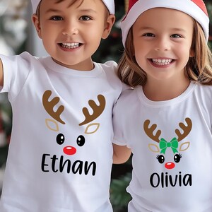 May include: Two white t-shirts with a reindeer face design. The shirt on the left says "Ethan" and the shirt on the right says "Olivia".