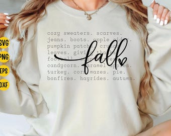Pode incluir: Camisola creme com a palavra "fall" em fonte cursiva. O design inclui uma lista de palavras com tema de outono como "suéteres aconchegantes", "campo de abóboras" e "passeios de feno".