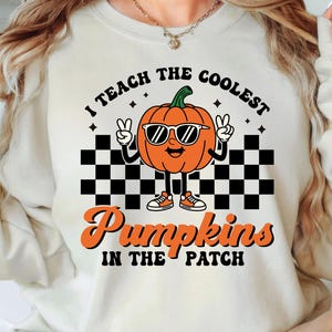 Könnte beinhalten: Cremefarbenes Sweatshirt mit dem Text "I teach the coolest pumpkins in the patch". Das Design zeigt einen Cartoon-Kürbis mit Sonnenbrille und Turnschuhen, mit einem schwarz-weißen Schachbrettmuster.