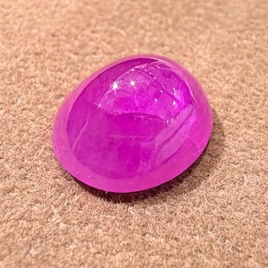 Op de afbeelding: Een gepolijste, ovaalvormige, magenta edelsteen. De edelsteen is glad en reflecterend, geplaatst op een bruin, getextureerd oppervlak. De belichting creëert highlights op het oppervlak van de edelsteen, waardoor de afgeronde vorm wordt benadrukt.