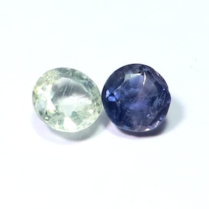 May include: Two gemstones on a white background. One is a pale green colour, and the other is a deep blue-purple. Both are oval-shaped with faceted surfaces, reflecting light. The gems appear to be polished and ready for jewellery.