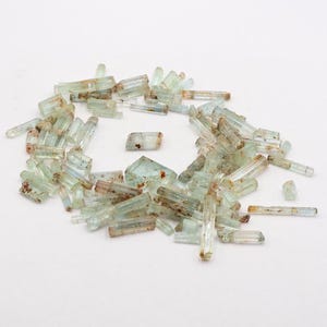 Peut inclure: Une collection de cristaux d'aigue-marine vert clair, certains avec des inclusions brunes, de différentes tailles et formes. Les cristaux sont disposés en cercle lâche sur un fond blanc.